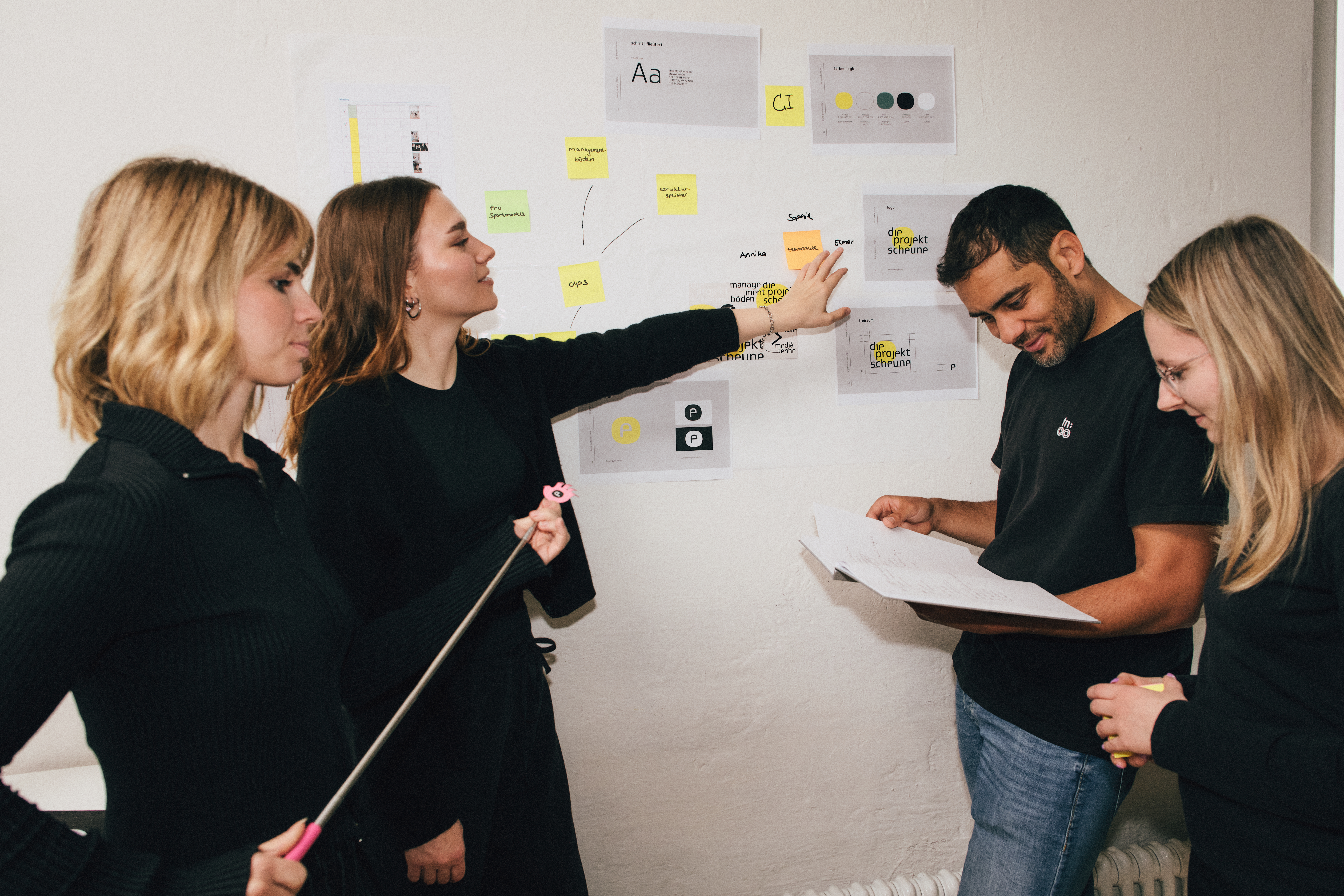 Team-die-projektscheune-2 Das Team steht gemeinsam am Whiteboard. Eine Person zeigt auf etwas, zwei andere schauen etwas in einem Buch nach.