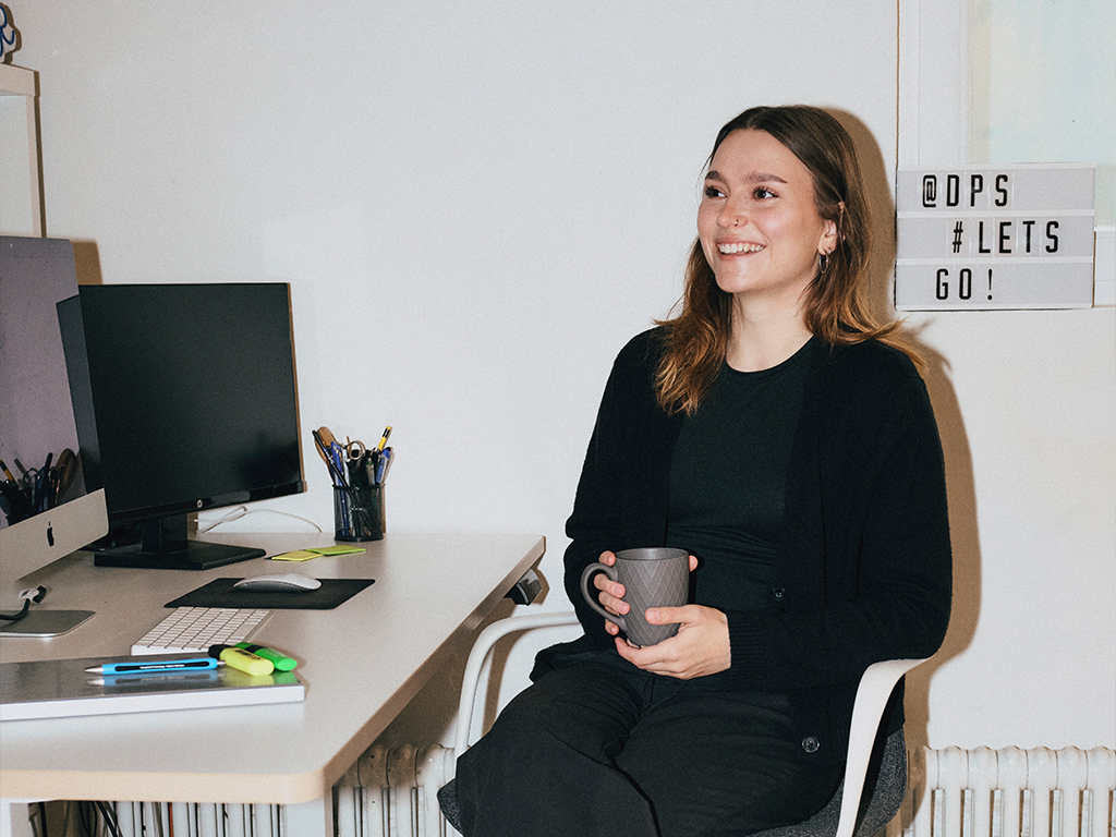 Annika-die-projektscheune Zu sehen ist Annika Maier, die Werkstudentin im Marketing und Social Media. Sie sitzt mit einer Tasse in der Hand am Schreibtisch.