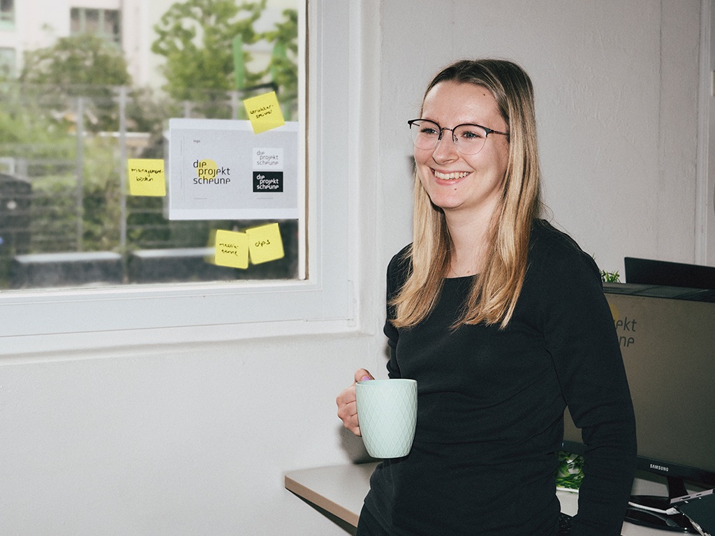 Sophie-die-projektscheune Zu sehen ist Sophie-Marie Schwab, die Assistenz der Geschäftsführung. Sie steht mit einer Kaffeetasse in der Hand am Schreibtisch.